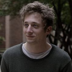 a young man with curly hair is looking at the camera while standing in front of a tree