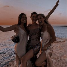 three beautiful women standing next to each other near the ocean at sunset or dawn with their arms in the air