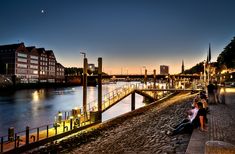 people are standing on the edge of a river at sunset or dawn with buildings in the background