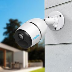 an outdoor security camera mounted on the side of a building with a house in the background