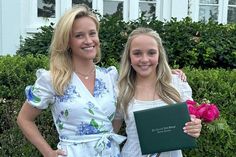 two women standing next to each other in front of bushes and flowers holding a book