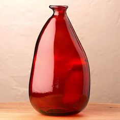 a red glass vase sitting on top of a wooden table next to a white wall