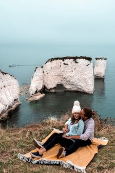 two people sitting on a blanket in front of the ocean with cliffs and water behind them