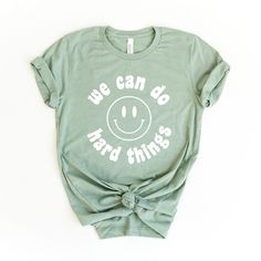 Looking for a cute versatile top to wear this summer? Make sure to grab one of our We can Do Hard Things Smiley Face tees! This soft and comfortable graphic tee is the perfect top for any outfit. It can be paired with biker shorts, jeans, or even a simple skirt/dress! This tee is true-to-size, so be sure to order your regular t-shirt size! If you are looking for a more oversized look, make sure to size up! Graphic Tees For Teachers, Inspirational Shirts Women, School Counselor Shirts, Tshirt Flatlay, Teacher Cricut, We Can Do Hard Things, Smiley Face Graphic, Smiley Face Tee, Slp Shirts