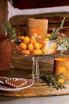 oranges in a glass bowl on a table next to candles and cookies with pine cones