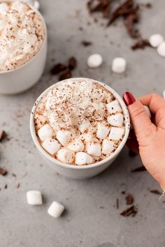 two cups of hot chocolate with marshmallows on the side, one being held up