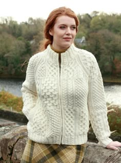 a woman wearing a white sweater and plaid skirt standing next to a stone wall with trees in the background