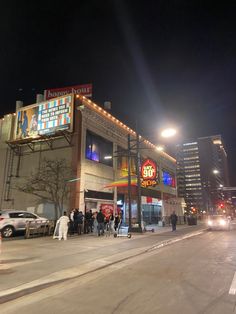 people are standing on the sidewalk outside of a building at night time with cars parked in front of it