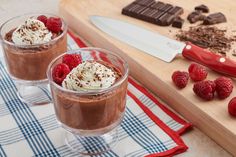 two glasses filled with chocolate pudding and raspberries next to a knife on a cutting board