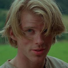 a young man with blonde hair and blue eyes looks at the camera while standing in front of a green field