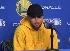 a man in a yellow hoodie sitting at a microphone with a basketball cap on