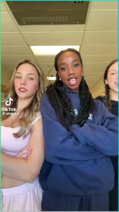 three girls are posing for the camera in an office building with their arms crossed and one girl is wearing a blue hoodie