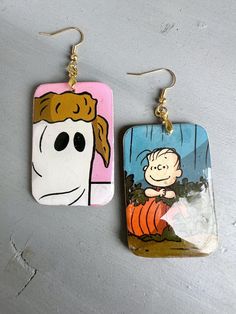 a pair of earrings with the image of charlie brown and pumpkins on them, hanging from gold ear wires