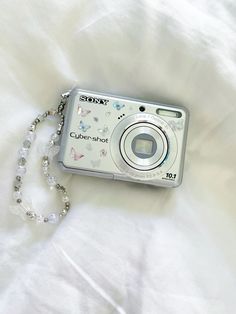 a white camera sitting on top of a bed next to a beaded chain necklace