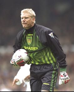 an older man holding a soccer ball and glove on his left hand while walking across a field