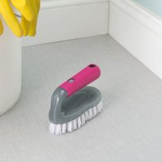 a pink and gray brush sitting on the floor next to a white vase with yellow gloves hanging from it