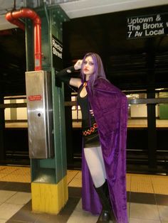 a woman dressed in purple and black standing next to a train station platform with her hand on her head