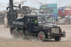 an old pick up truck driving down a dirt road with other trucks in the background