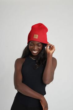 a woman wearing a red beanie smiles while holding her hair in one hand and smiling at the camera