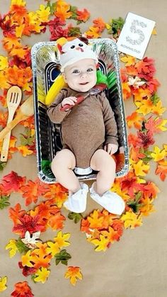 a baby is sitting in a tray surrounded by fall leaves and other autumn flowers, with a hat on it's head