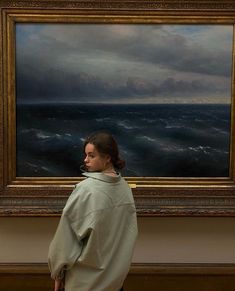 a woman standing in front of a painting looking at the ocean with storm clouds overhead