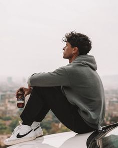 a man sitting on top of a car holding a beer in his right hand and looking off into the distance