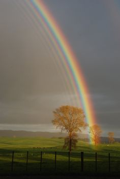 two rainbows in the sky over a green field with trees and a black fence