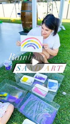 a woman sitting on the grass holding up a rainbow art piece next to her child