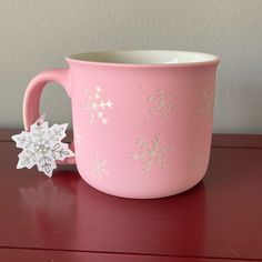 a pink coffee cup with white snowflakes on the side and a paper snowflake attached to it