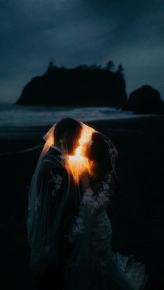 a bride and groom standing under an umbrella on the beach at night with light coming from behind them