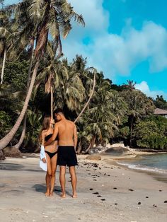 two people standing on the beach with palm trees in the background and one person wearing a black swimsuit