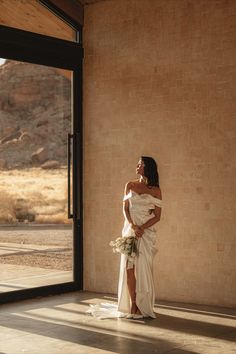 a woman standing in front of a window wearing a white dress and holding a bouquet