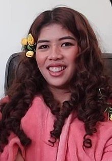 a woman with long curly hair and flowers in her hair smiles at the camera while sitting on a chair