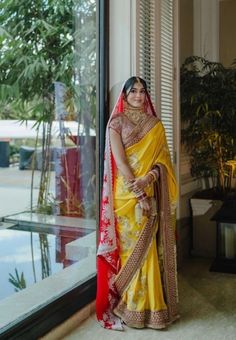 a woman in a yellow and red sari standing next to a large window with a potted plant behind her