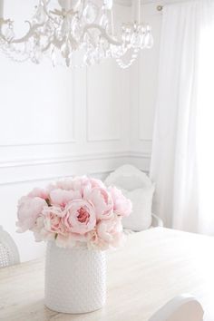 a white vase filled with pink flowers sitting on top of a wooden table next to a chandelier