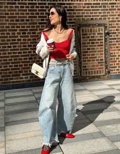 a woman in red shirt and jeans standing next to brick wall with her hand on her hip