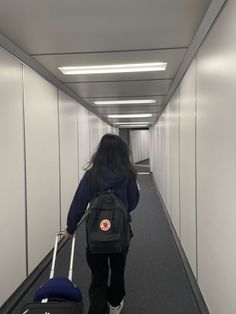 a woman with a backpack and suitcase walking down a long hallway holding a rolling luggage bag