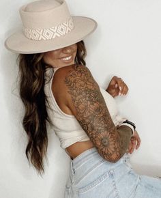 a woman wearing a white hat with tattoos on her arm and arms, leaning against a wall