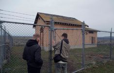 two men standing next to a fence with a building in the back ground behind them