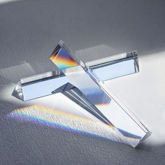 a cross made out of glass sitting on top of a white surface with a rainbow colored light coming from behind it