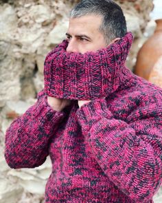 a man wearing a pink and black sweater with a cowl on his head standing in front of a stone wall