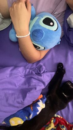 a woman laying in bed next to a black cat and a stuffed animal on her stomach