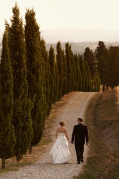 a bride and groom walking down a dirt road in front of tall evergreen trees at sunset