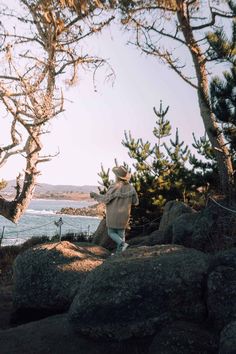 a man standing on top of a large rock next to the ocean in front of some trees