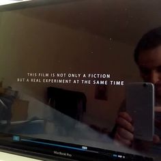 a man looking at his cell phone in front of a television screen that says, this film is not only a fiction but a real experiment at the same time