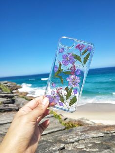 a hand holding up a phone case with flowers on it near the beach and ocean