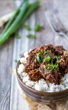 a bowl filled with rice and meat on top of a wooden table next to green onions