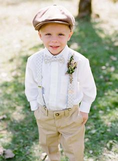 a young boy wearing a hat and bow tie standing in the grass with his hands on his hips