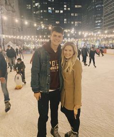 a man and woman posing for a photo on an ice rink at night with people skating in the background