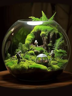 a fish bowl filled with green plants and people in the background on top of a wooden table